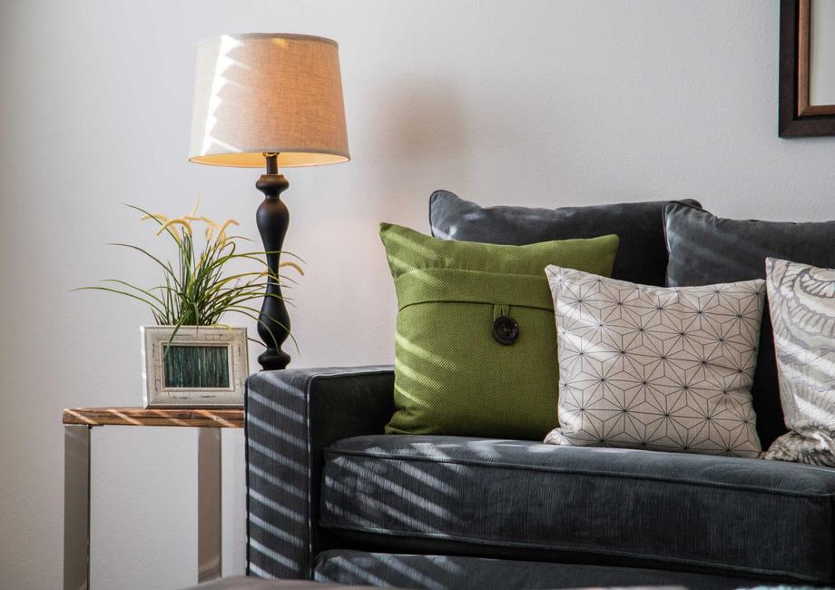 Dark blue sofa that has green and white pillows, framed picture hanging on the wall and lamp with beige shade in the corner.