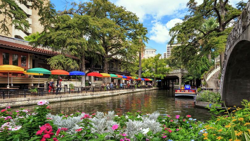 San Antonio, Texas Riverwalk showing blooming pink, white, and yellow flowers, multi-color sunshades and tables lining the river, and overgrown trees