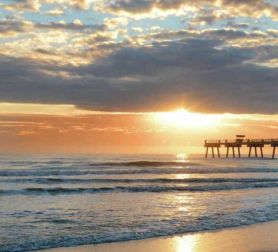 Jacksonville, Florida beach sunrise with pier extended far into the ocean, rolling waves, and sunlight reflecting off the wet sand