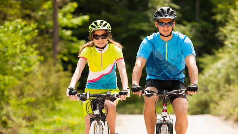 Stock image of a young couple wearing cycling clothing riding mountain bikes on a gravel path with large evergreen trees in the back ground.