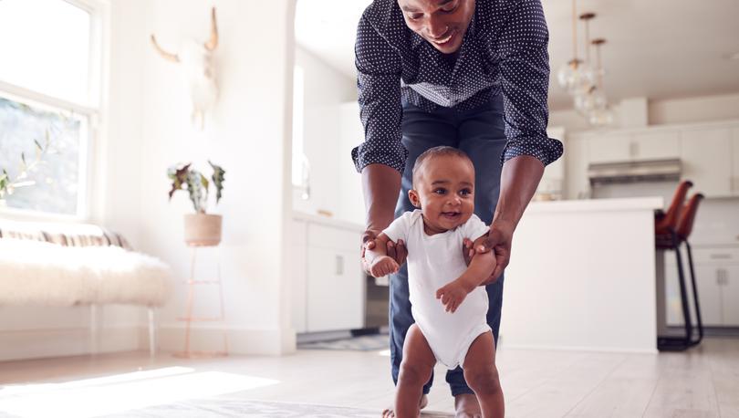 Dad smiling while helping his baby take its first steps.