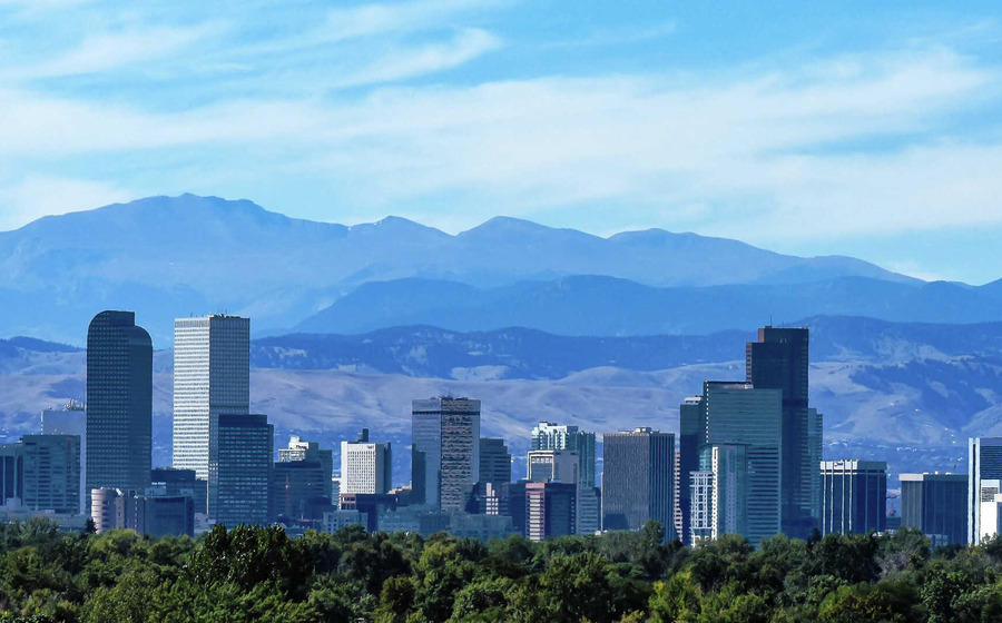 Denver, Colorado city skyline with multiple skyscrapers, trees in the foreground, and the Rocky Mountains in the distance