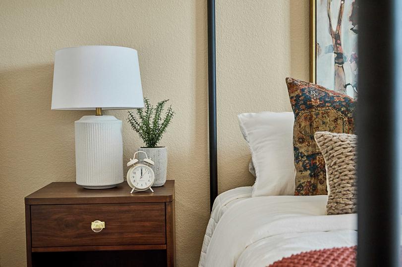 Detail of bed with iron posts, wooden side table with lamp, white sheets and warm natural throw pillows, blanket, nightstand with clock and plant and artwork at head of bed.