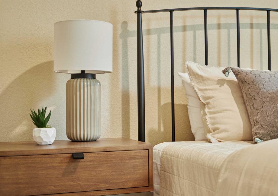 Staged bedroom photo showing a bed with pillows and a bedside table with a lamp and plant.