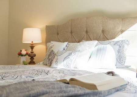 This bedroom is staged with a sandy color button tufted headboard, open book on top of a bluish comforter with white pillows on the bed, and a lit lamp with beige shade