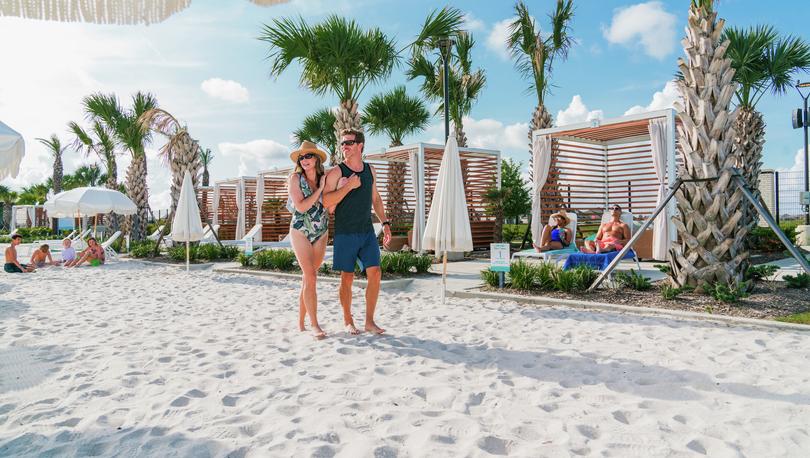 Couple in swimsuits walks in white sand along the private cabana area with palm trees as children play in the sand behind them.