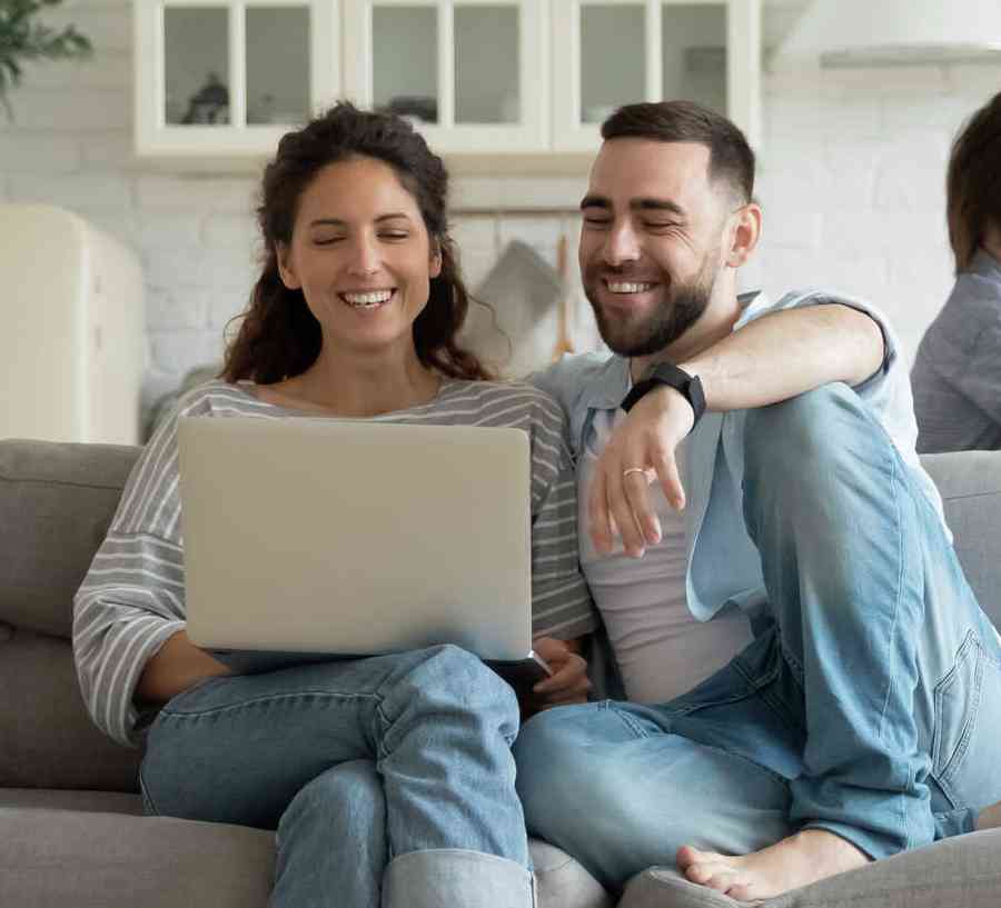 Couple sitting on a gray couch looking at a laptop, while two children play in the room.