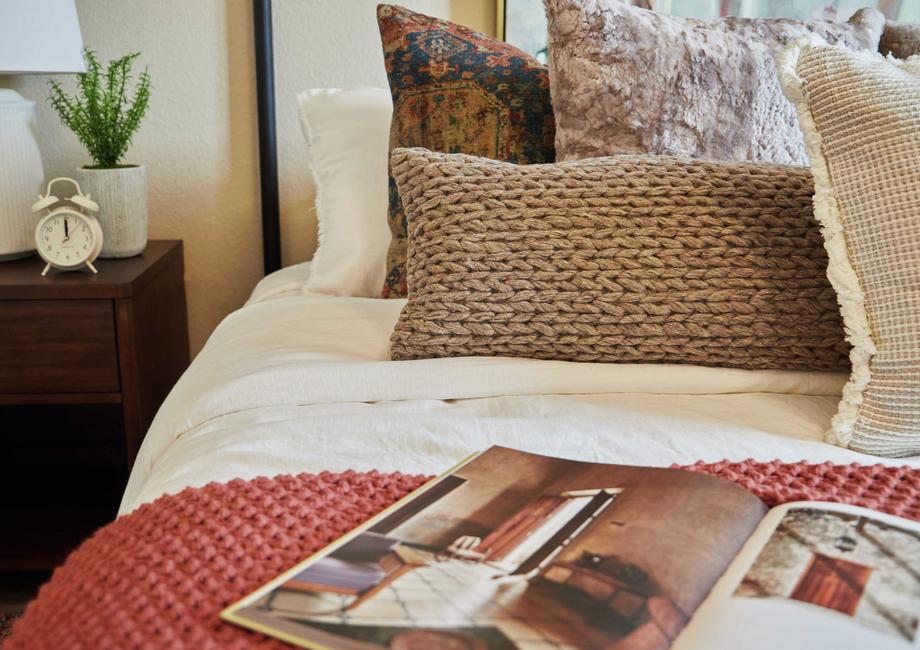 This bedroom has been staged with gorgeous white and rust colored bedding, an abundance of accent pillows, and a wood end table.