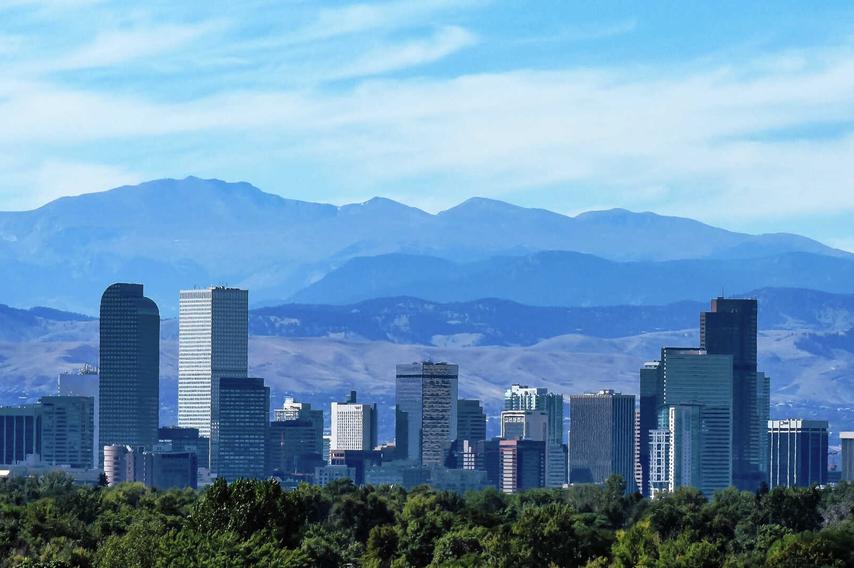 Denver, Colorado city skyline with multiple skyscrapers, trees in the foreground, and the Rocky Mountains in the distance