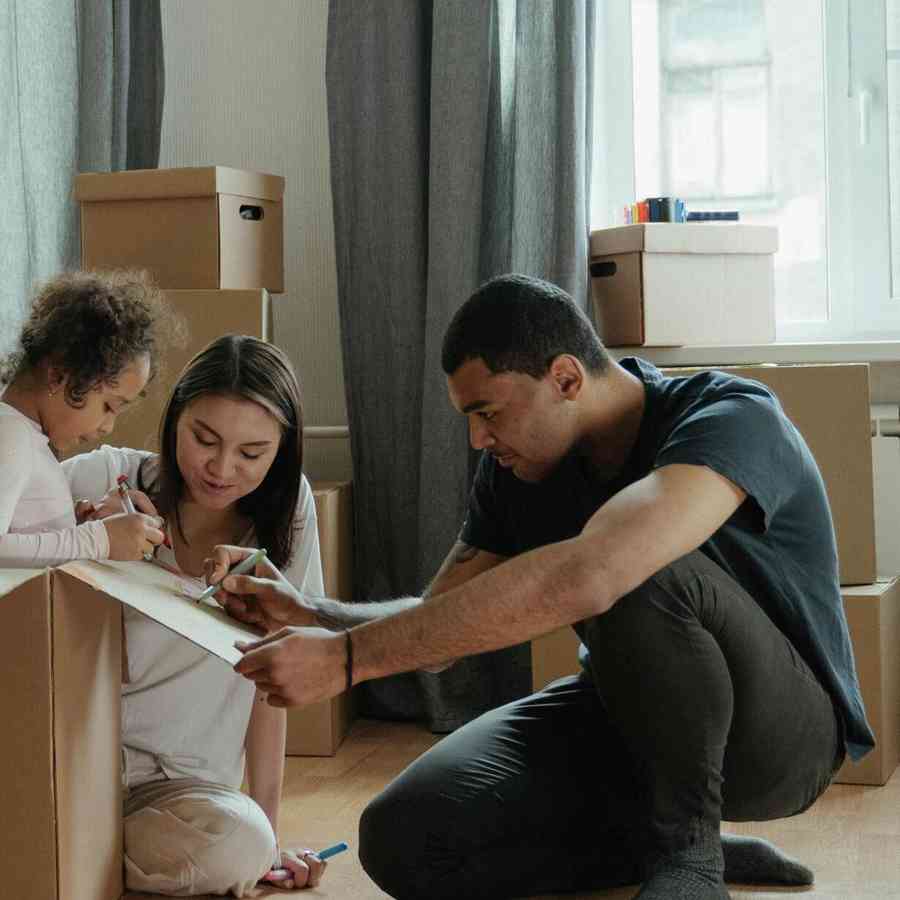 A young family sits on the floor drawing on a box and is surrounded by moving boxes.