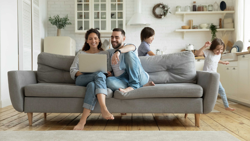 Couple sitting on a gray couch looking at a laptop, while two children play in the room.