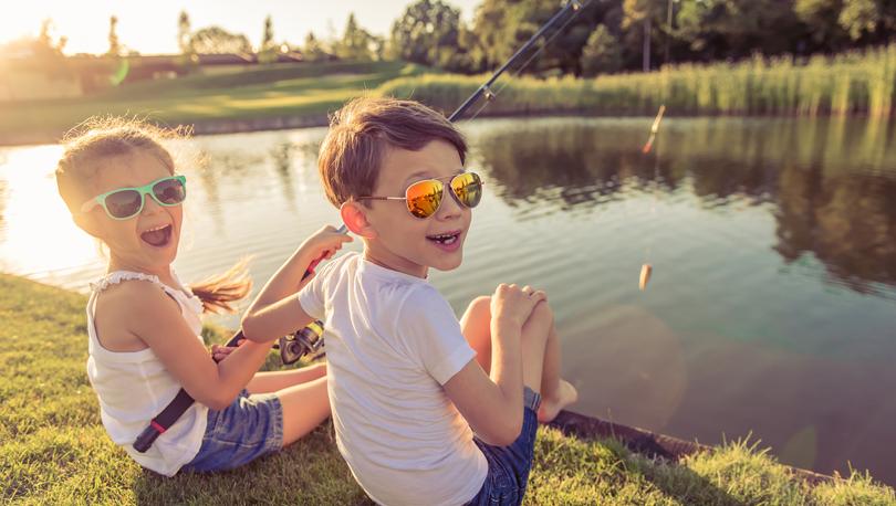 Funny stylish little boy and girl in sun glasses are looking at camera and smiling while catching fish in the pond using a fishing rod, sitting on the ground