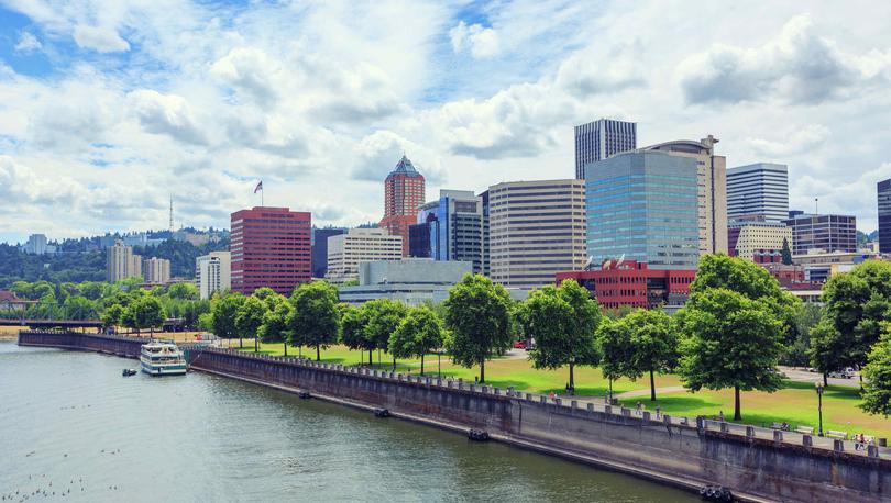 Portland, Oregon skyline taken from the river showing a docked boat, trees and grass on the bank, and office buildings