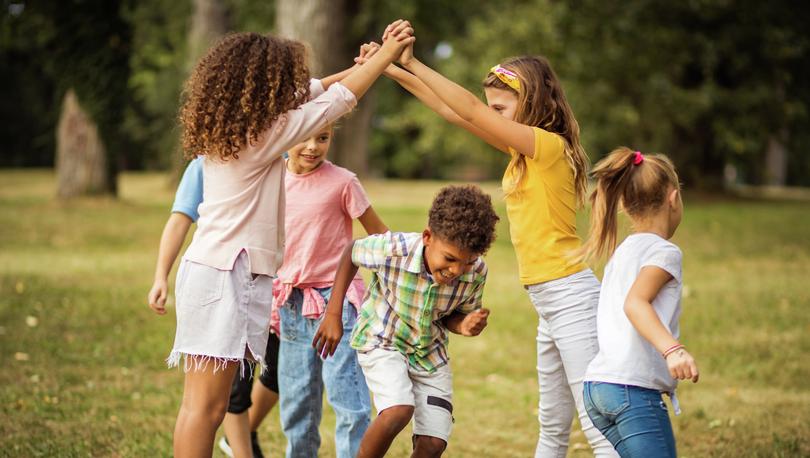 Group of young children playing outdoors at a park in the grass with trees in background.