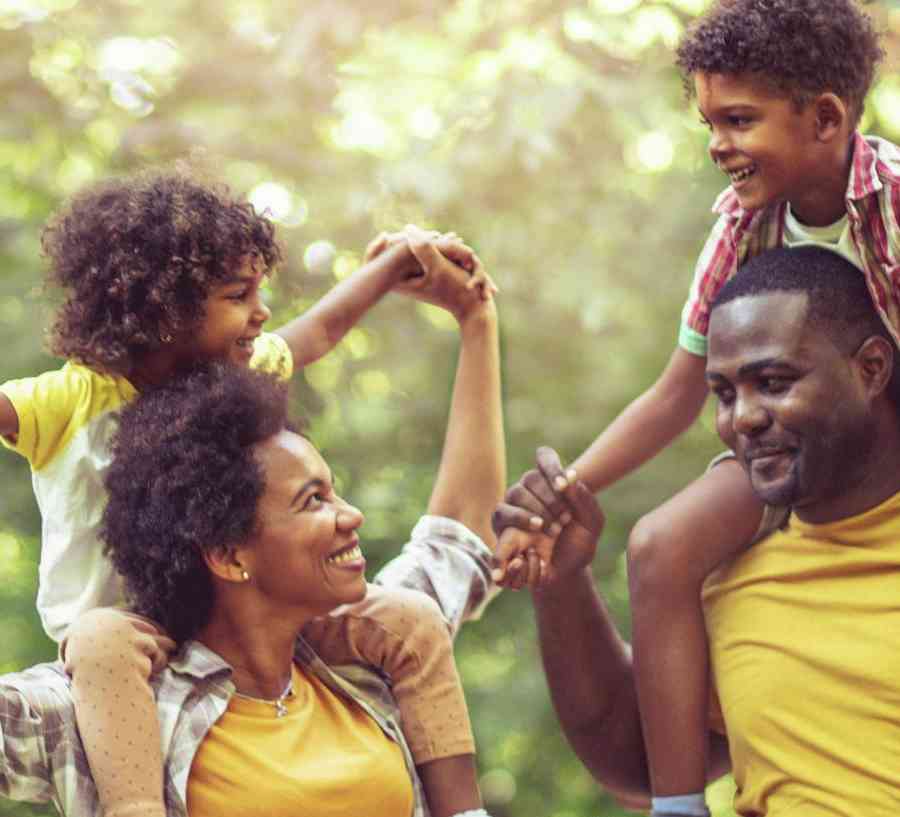 Family walking through park on sunny day with tall trees in background and kids on parents