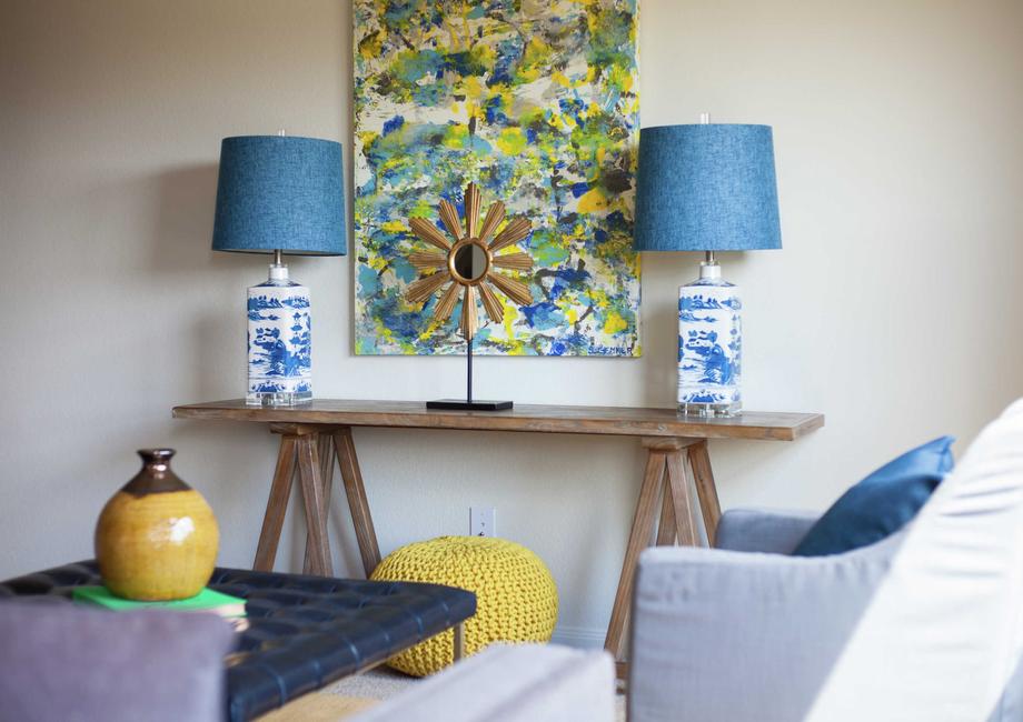 Staged living room with two decorative lamps with blue shades on a wooden table, painting hanging on the wall and a yellow vase on a navy coffee table.