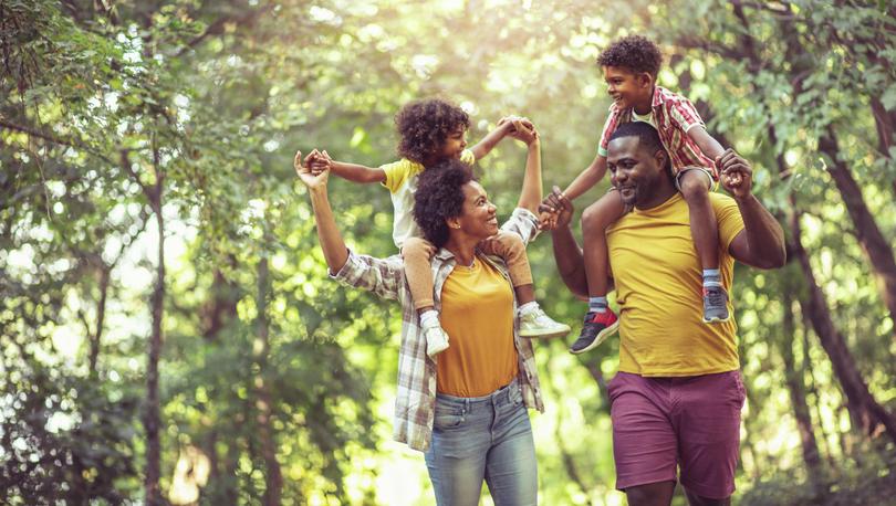 Family walking through park on sunny day with tall trees in background and kids on parents' shoulders.