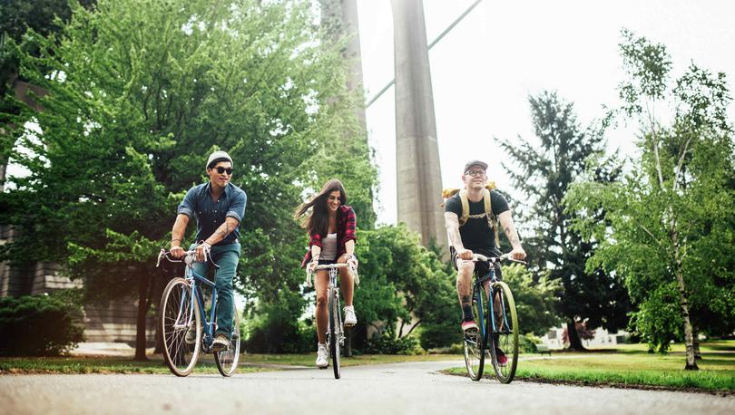 Two young men and one woman riding bikes through a Portland, OR park with green trees and bridge in the background